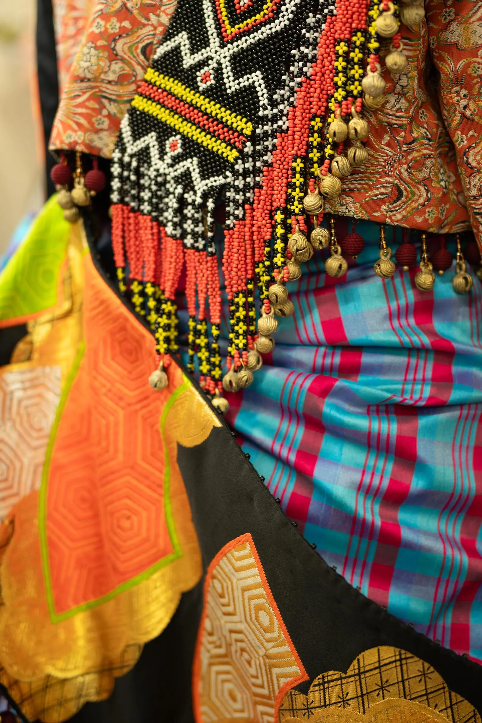 Close-up of bead-embroidered fabric with geometric patterns in red, black, yellow, and white, plus dangling tassels and patterned textiles.