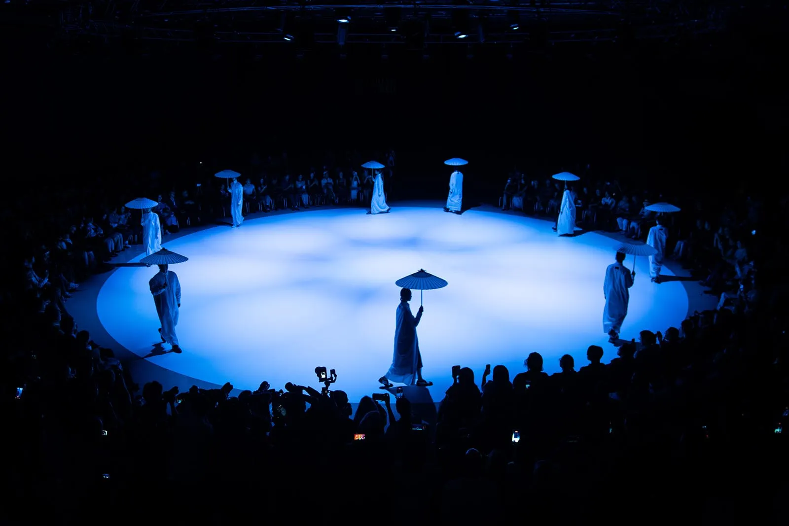 Performers in white robes with blue umbrellas circle a lit blue stage, audience seated around in darkness, blue spotlight creates round performance space