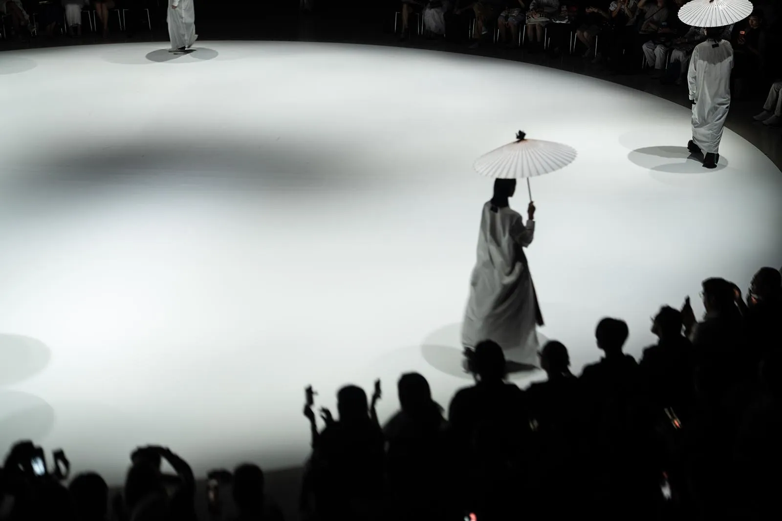 Two models in white robes walk a circular white runway, each holding a white parasol, as spectators watch from the dark audience.