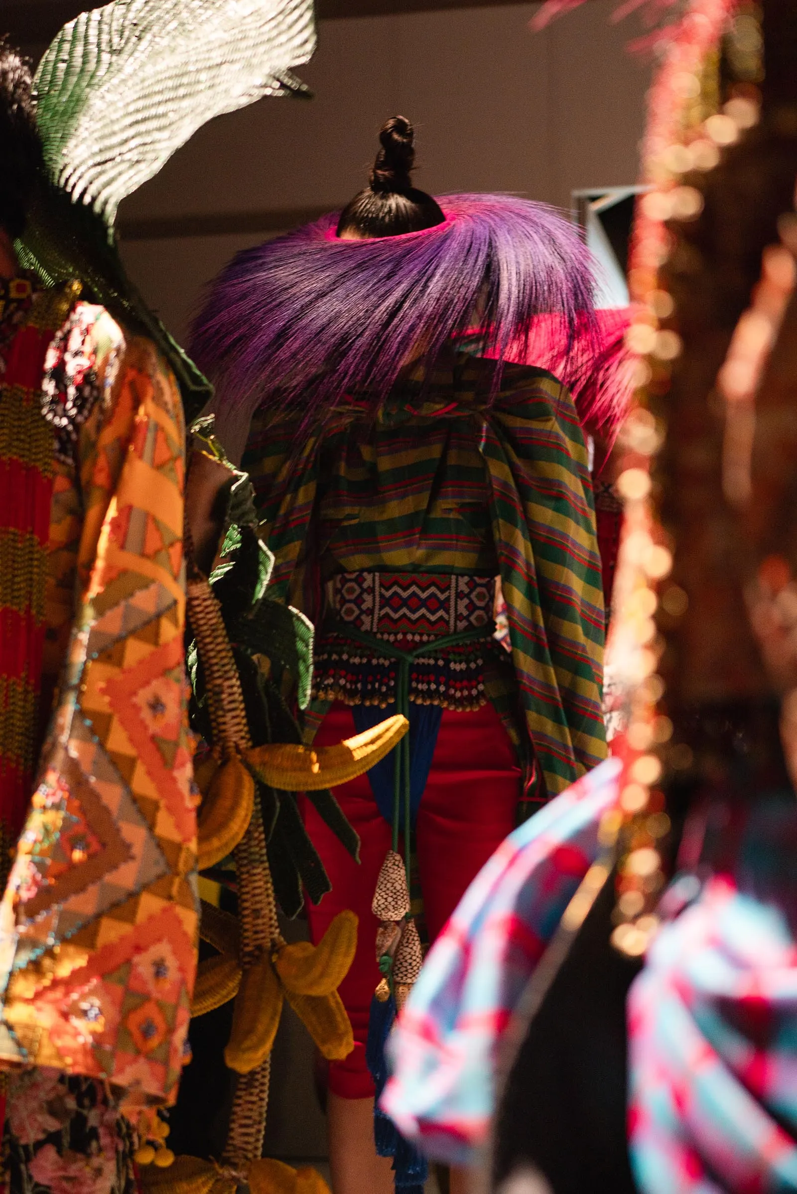 Person with vibrant purple-pink hair in a colorful traditional outfit seen from behind among decorative fabrics and tassels at a festival or market.