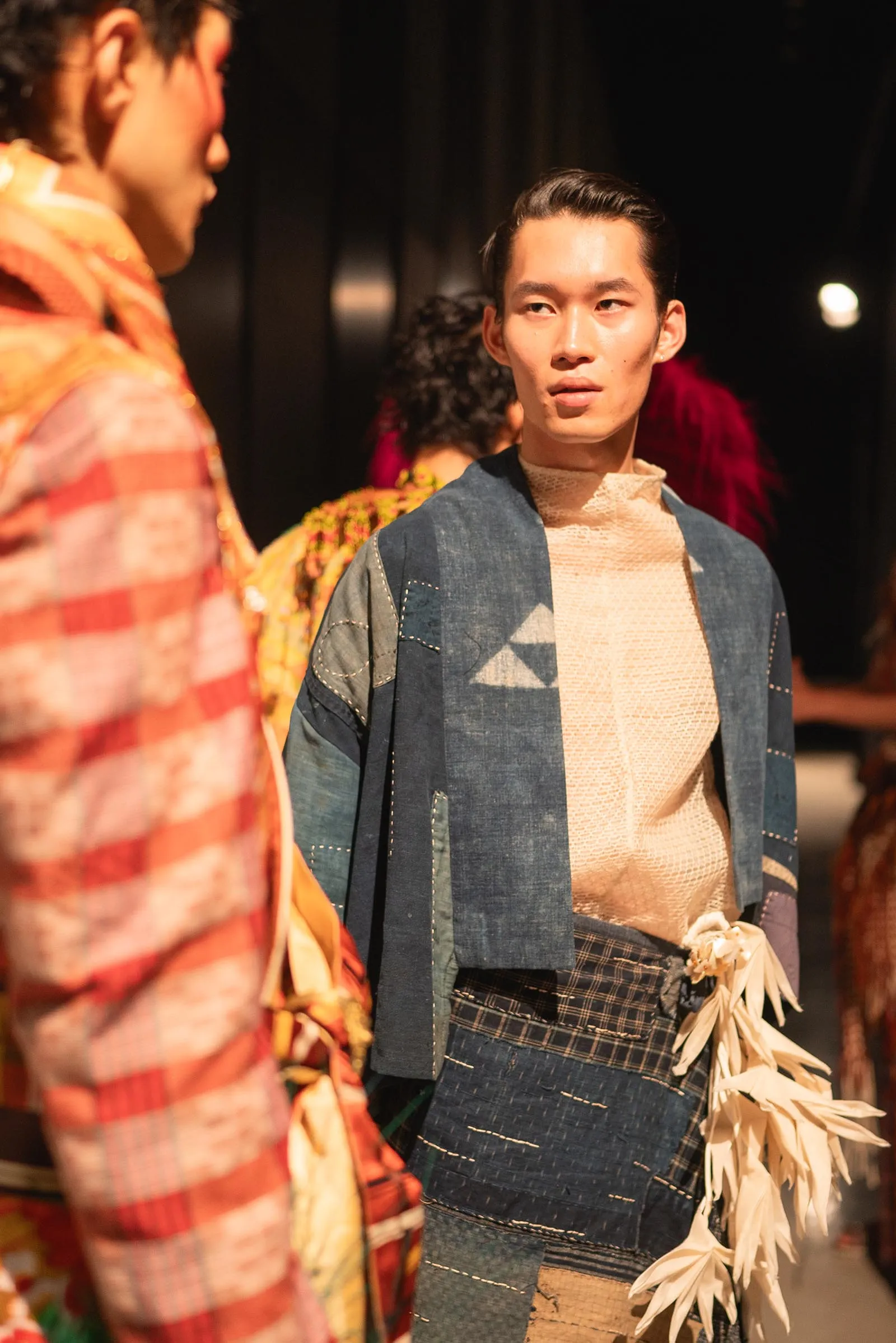 A man wearing a denim jacket and beige top stands in a backstage area, looking toward another model off-camera.