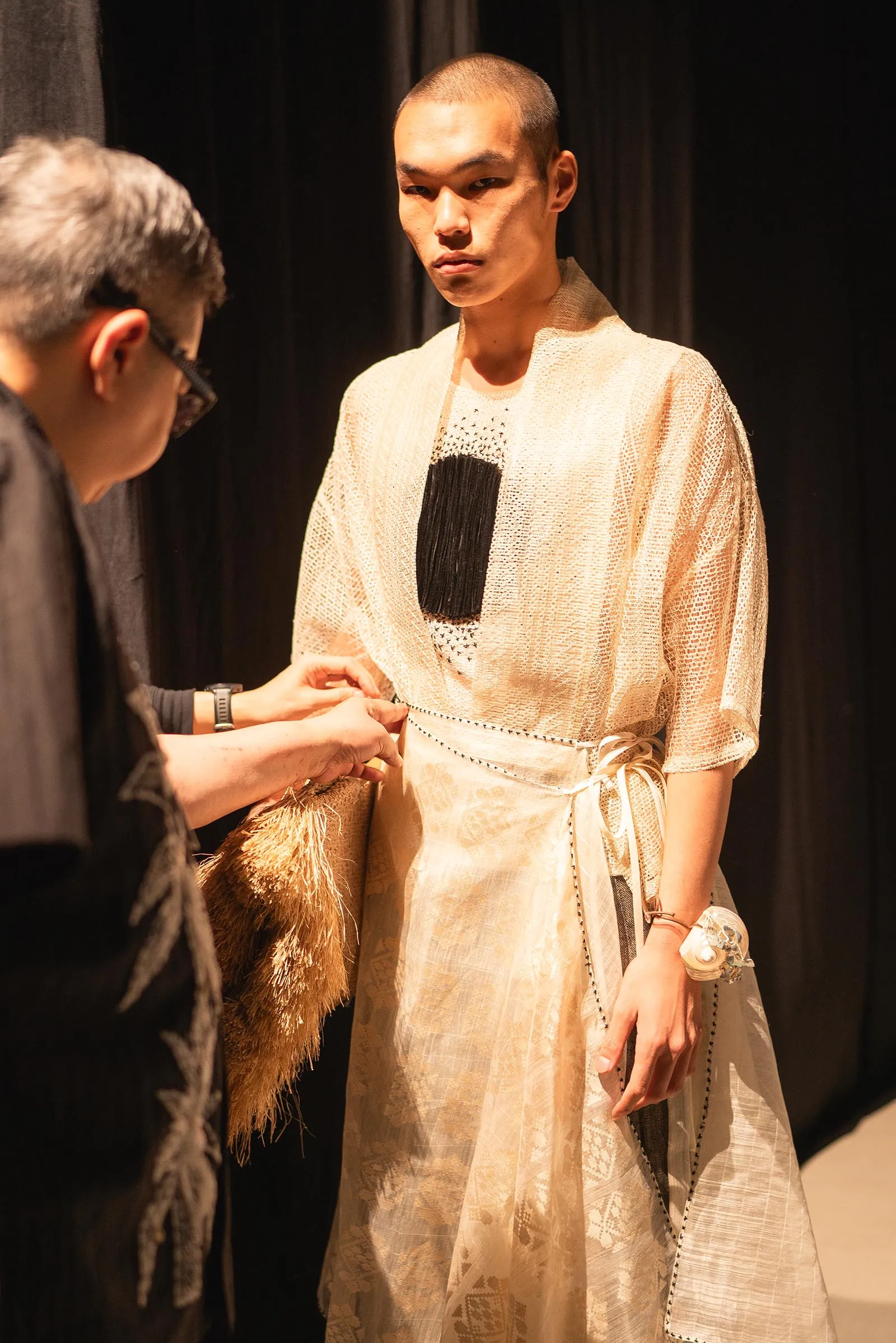 Model in a cream textured robe with a black chest panel, standing on stage as a stylist adjusts her outfit.