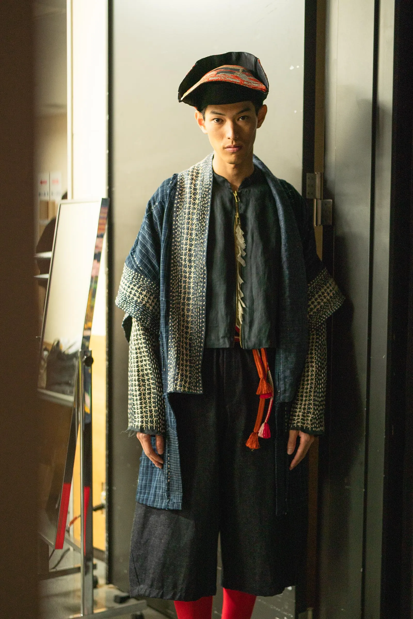 Person in a blue patterned traditional robe with a black hat stands in an elevator, facing the camera.